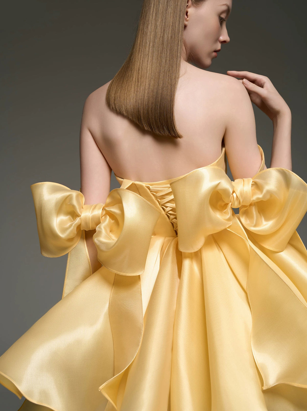 A woman shown from the back wearing a strapless formal dress made of shiny pale yellow satin, with oversized bow details at the shoulders and a large bow across the back. The photo is set against a dark gray studio background.