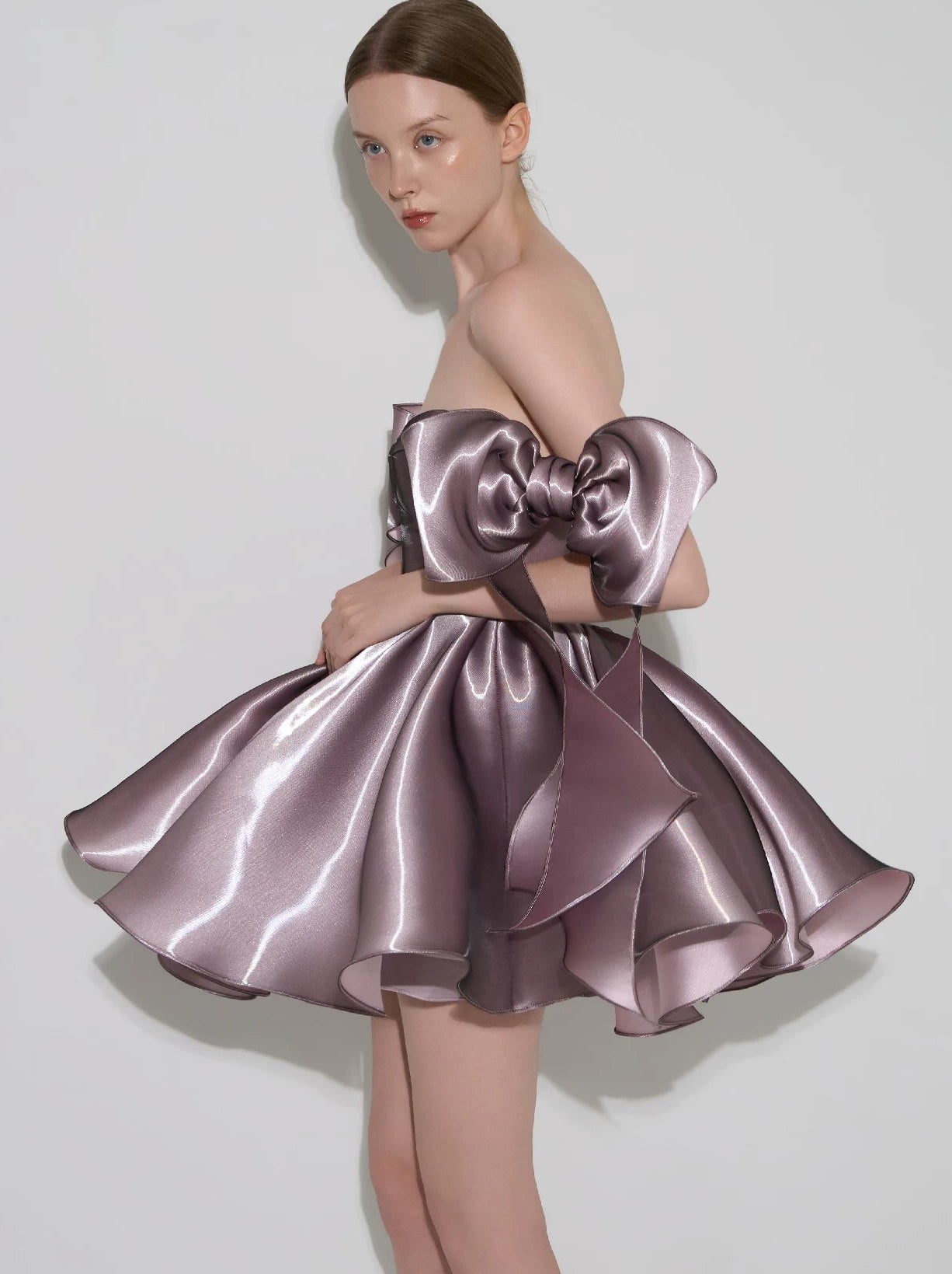 A model wearing a strapless, off-the-shoulder mini dress made of shiny mauve-purple satin, featuring a large oversized bow at the bodice and a ruffled skirt, set against a plain light gray studio background.