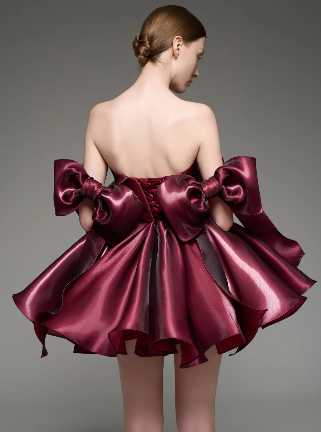 A model shown from the back wearing a strapless burgundy satin mini dress with oversized bow details on the shoulders and a ruffled skirt, set against a solid gray studio background.