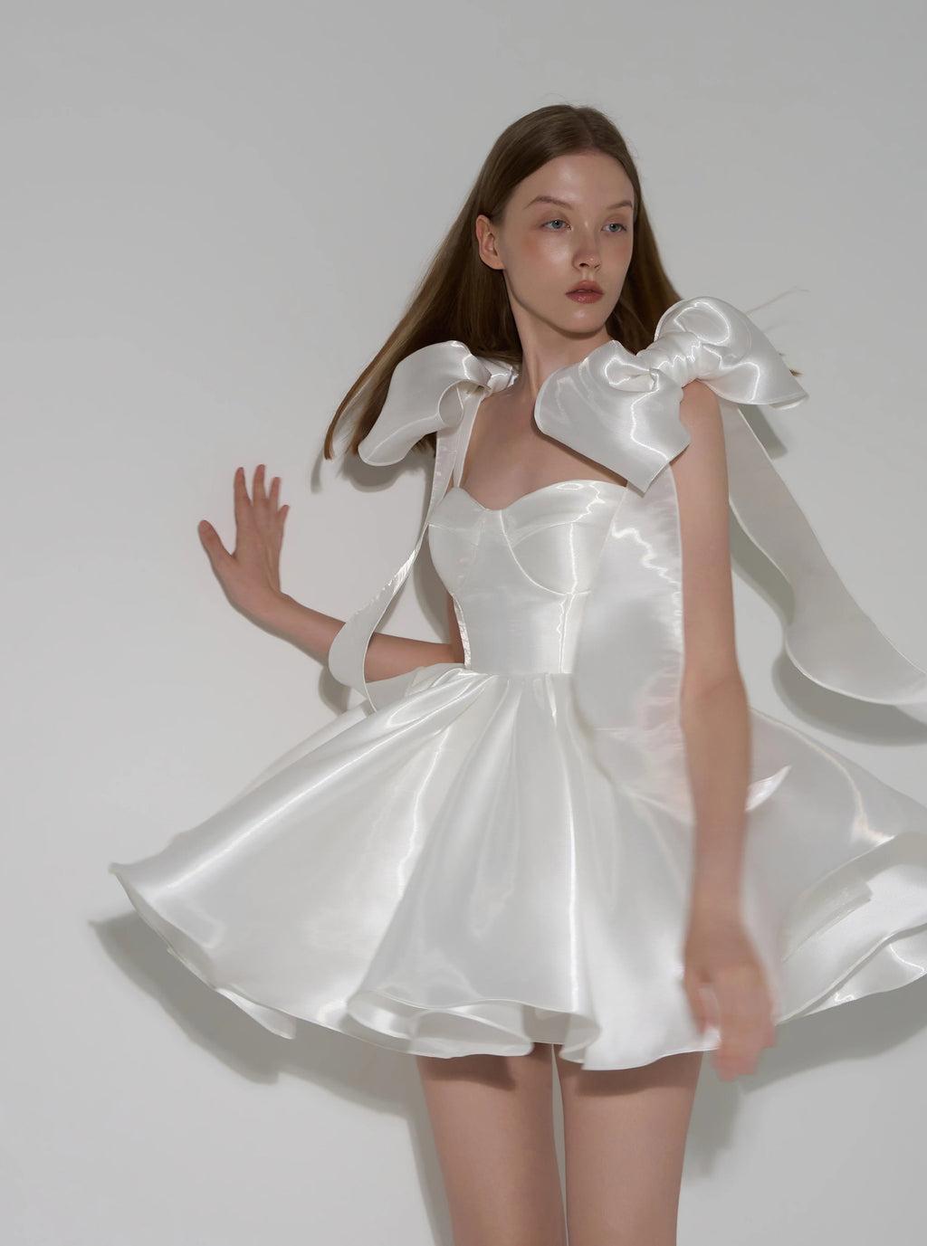A model wearing a white satin dress with dramatic oversized ruffled shoulder details and a flared skirt, posed against a plain light gray studio background.