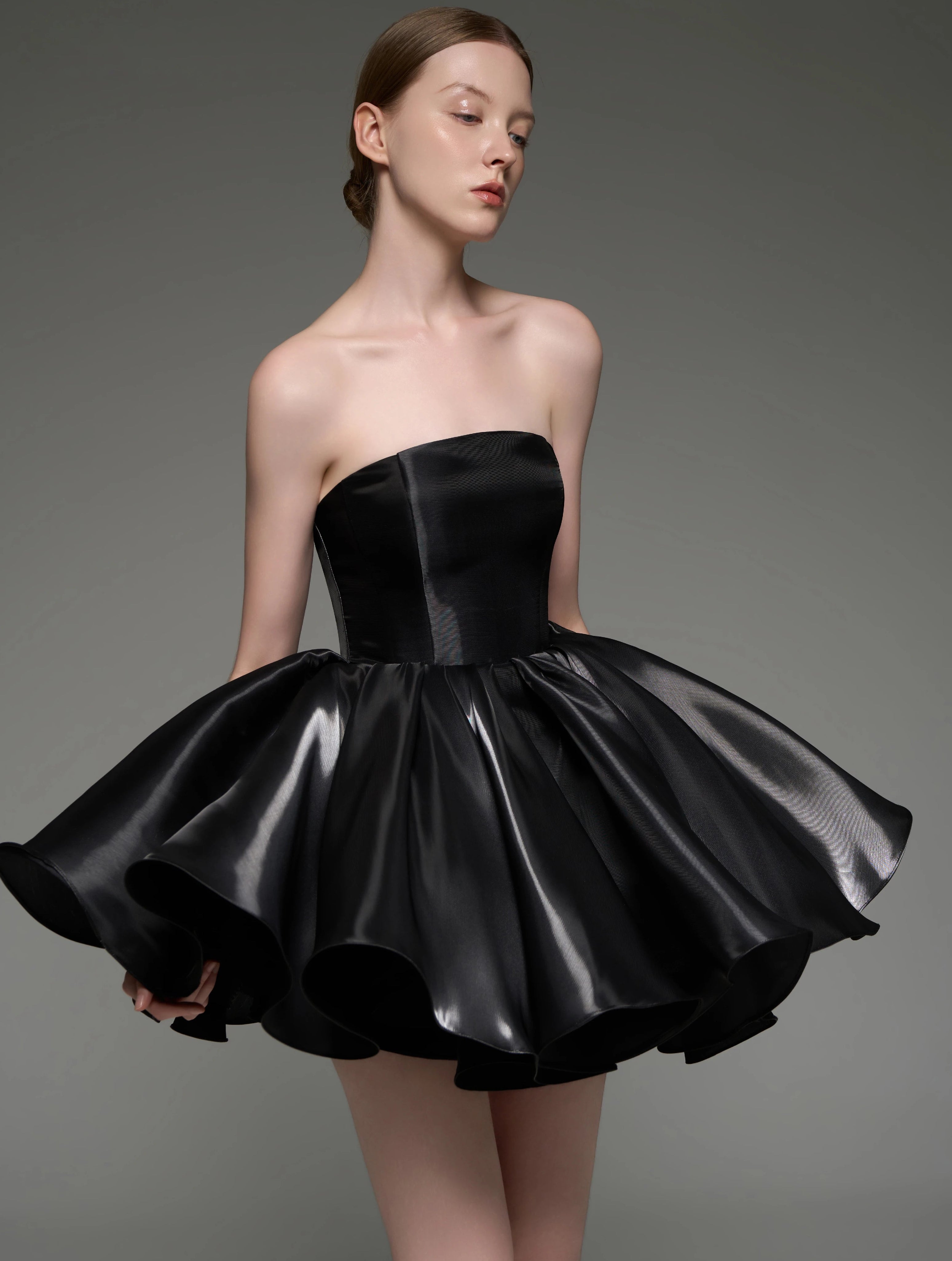 A woman modeling a strapless black formal dress with a structured corset-style bodice and a voluminous layered tulle skirt, photographed against a plain gray studio background.