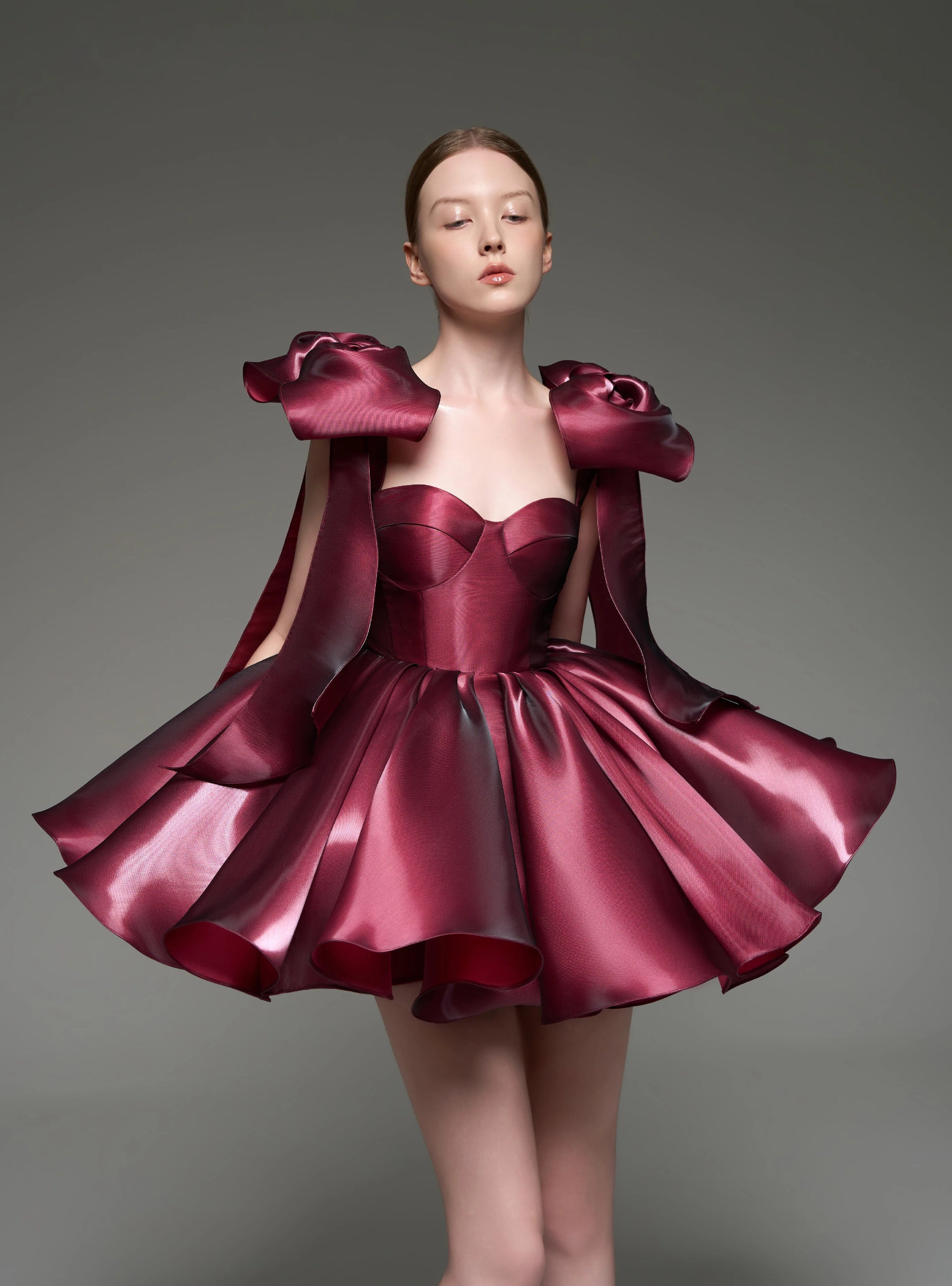 A model wearing a custom formal dress made of shiny, pleated burgundy satin fabric, featuring dramatic shoulder bows and a voluminous skirt, set against a solid gray studio background.