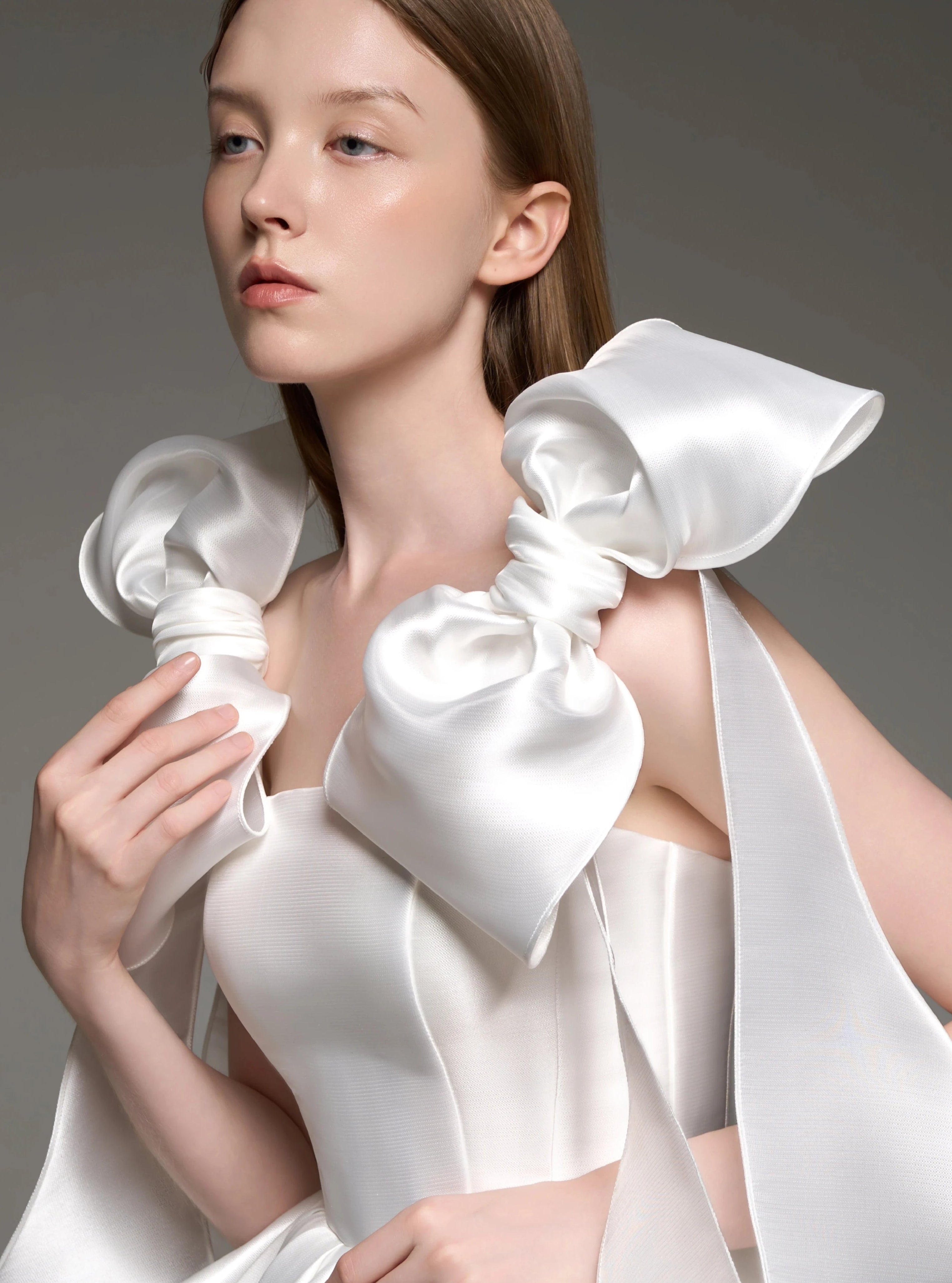 A model wearing a white satin formal dress with dramatic oversized bow details on the shoulders, photographed against a neutral gray studio background.