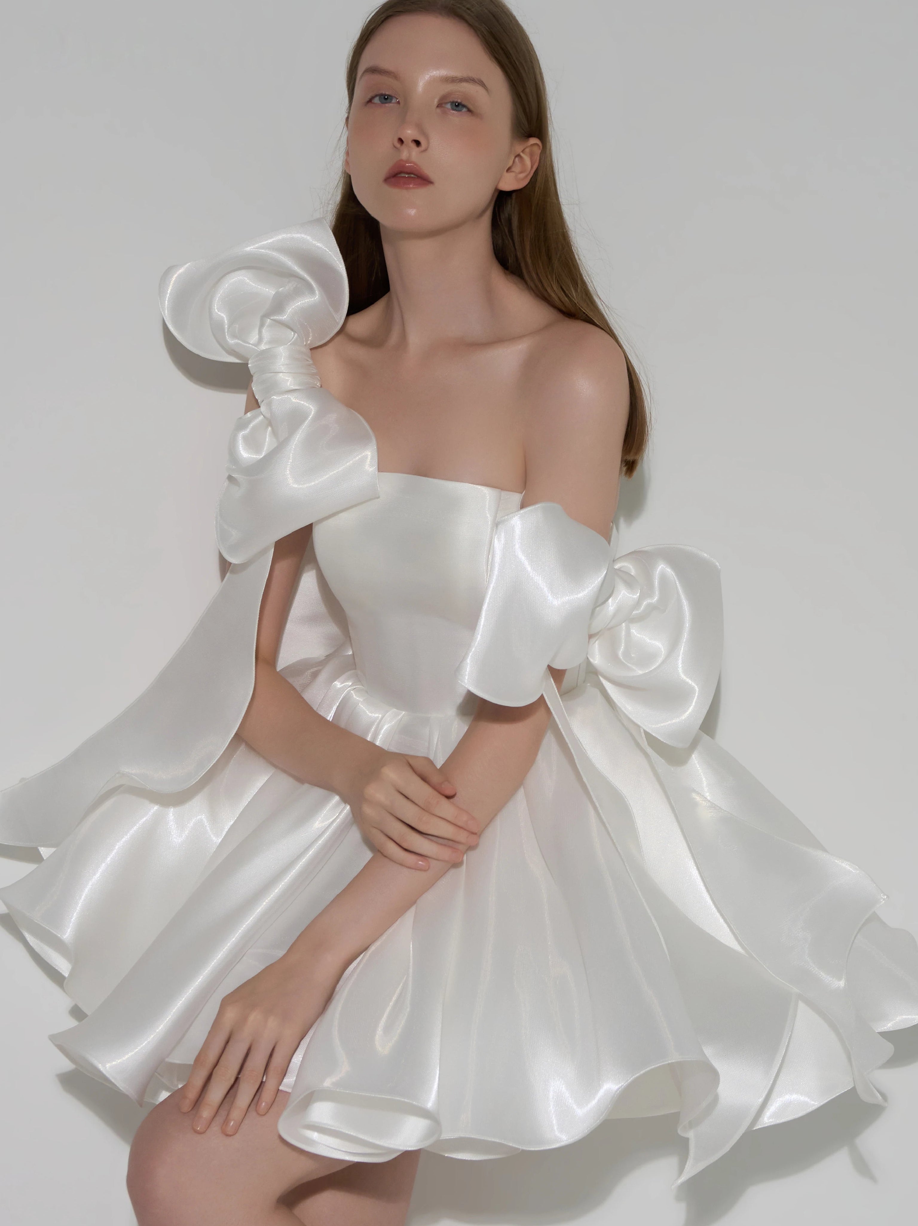 A model wearing a white satin off-the-shoulder mini dress with dramatic oversized bow details on the shoulders and a flared skirt, posed against a light gray studio background.