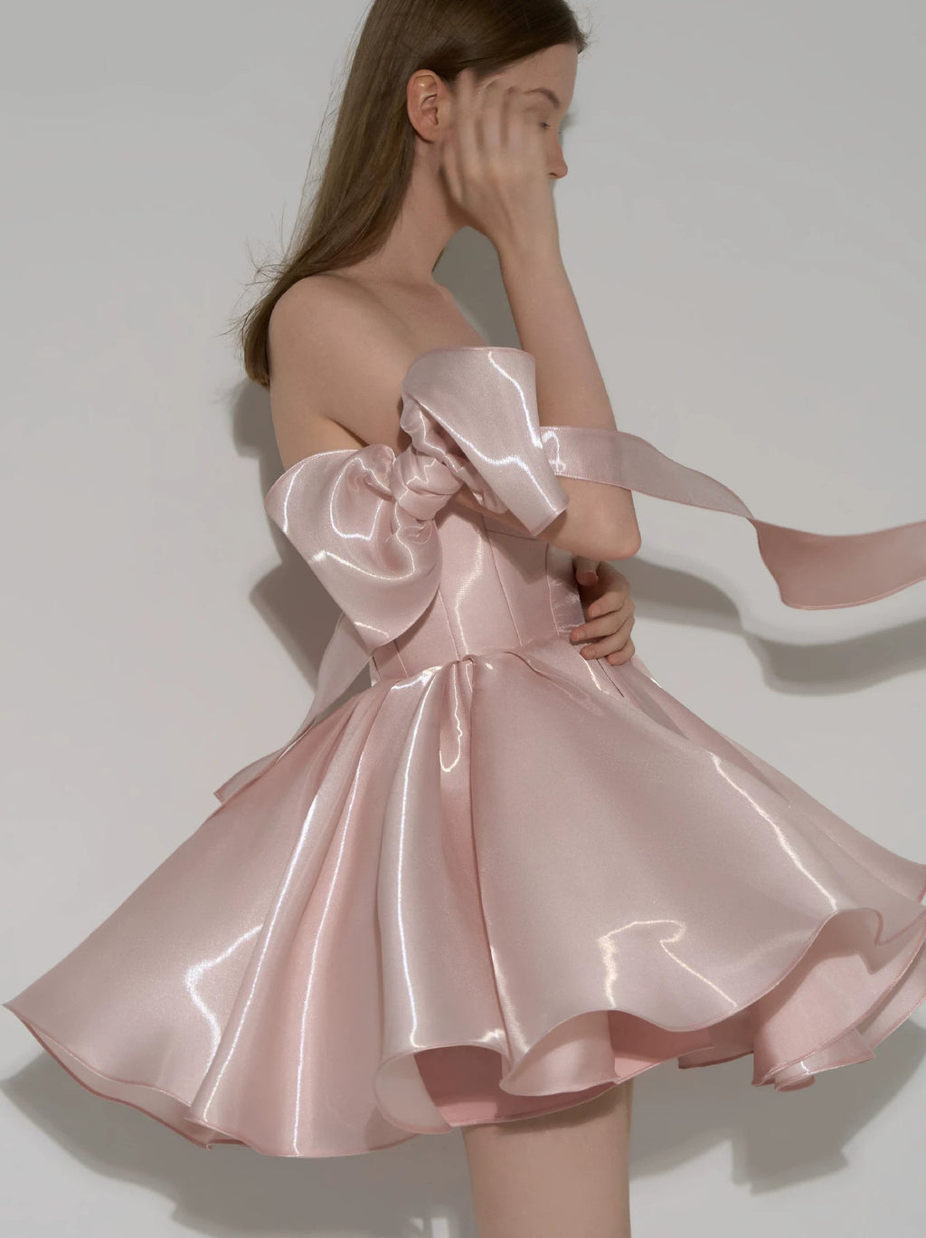 A model wearing a shiny, off-the-shoulder light pink satin dress with a voluminous skirt and a long flowing ribbon detail, posed against a neutral gray studio background.