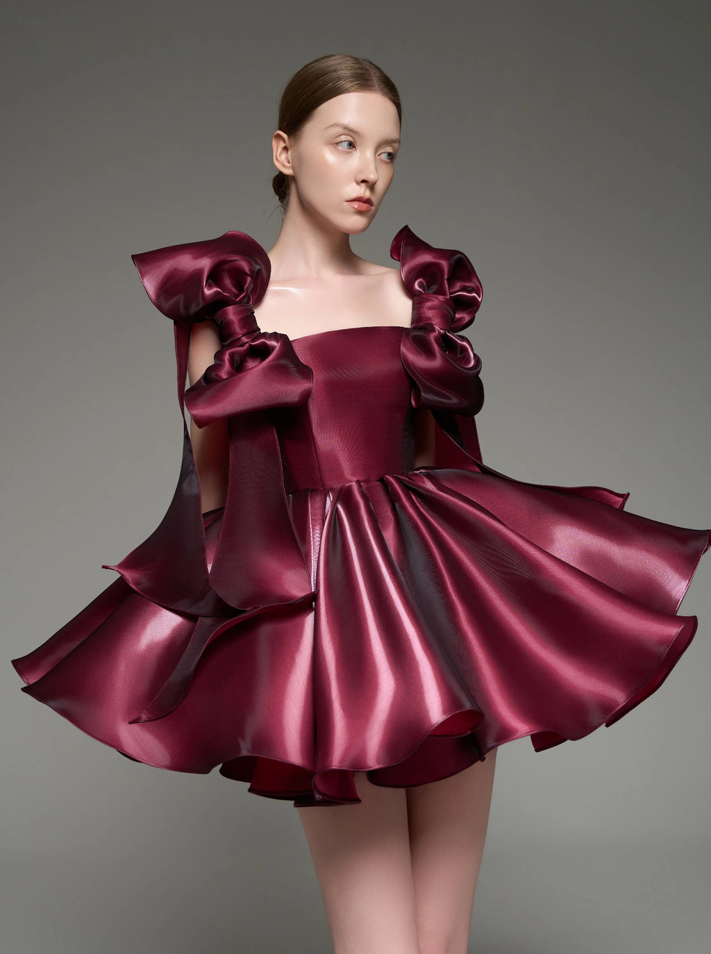 A model wearing a custom formal dress made of shiny deep burgundy satin, featuring dramatic oversized shoulder bows and a flared skirt, set against a solid gray studio background.