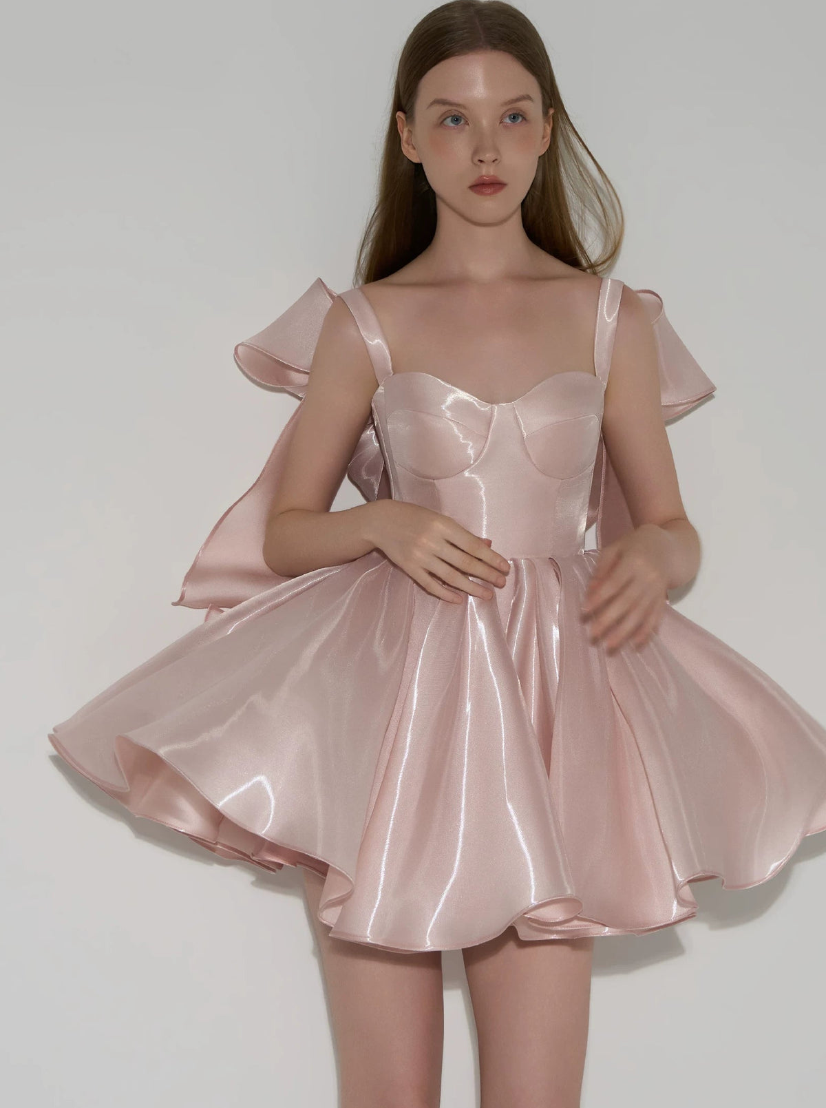 A model wearing a light pink satin formal dress with a structured sweetheart bodice, ruffled cap sleeves, and a flared skirt, shown against a plain light gray studio background.