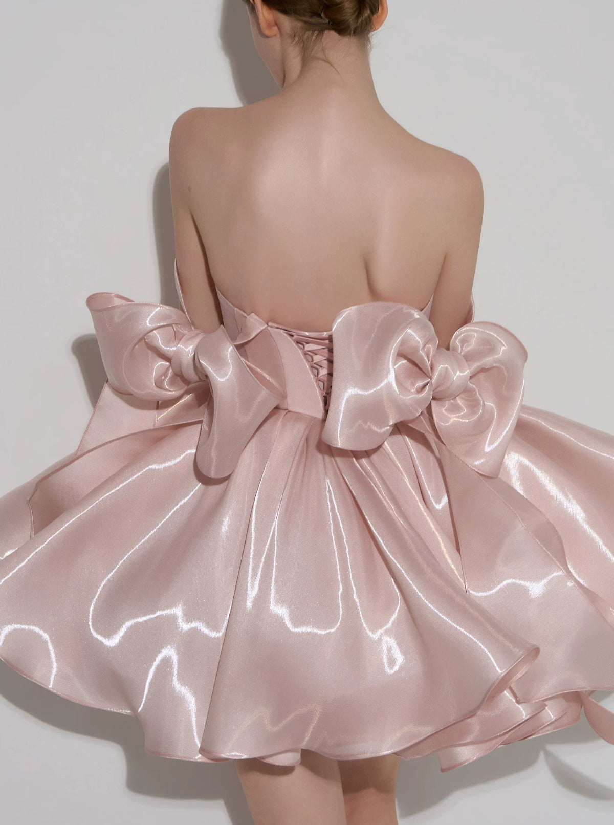 A model shown from the back wearing a strapless formal dress made of shiny light pink satin, featuring a large bow at the back and a flared skirt. Small text at the top reads 'Customized formal dress'.