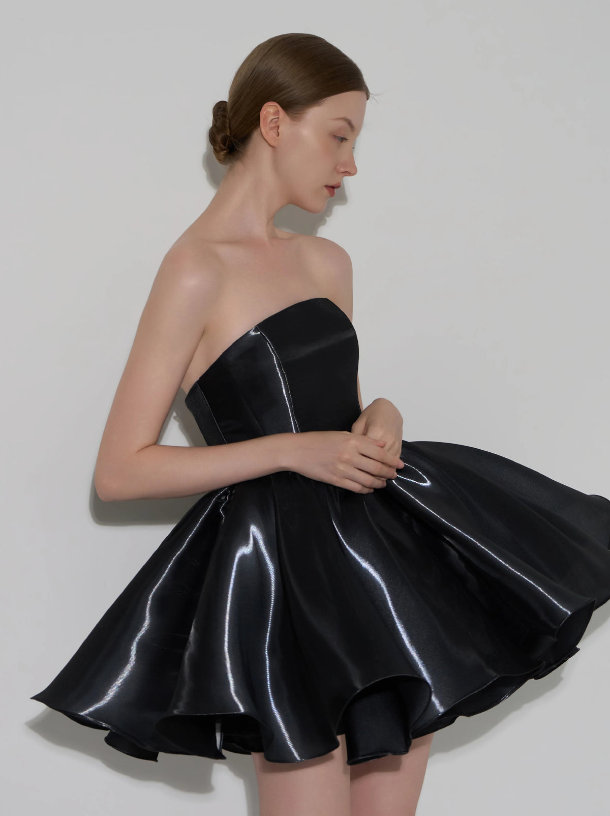 A woman modeling a strapless black formal dress with a structured corset-style bodice and a flared skirt, posed against a plain light gray studio background.