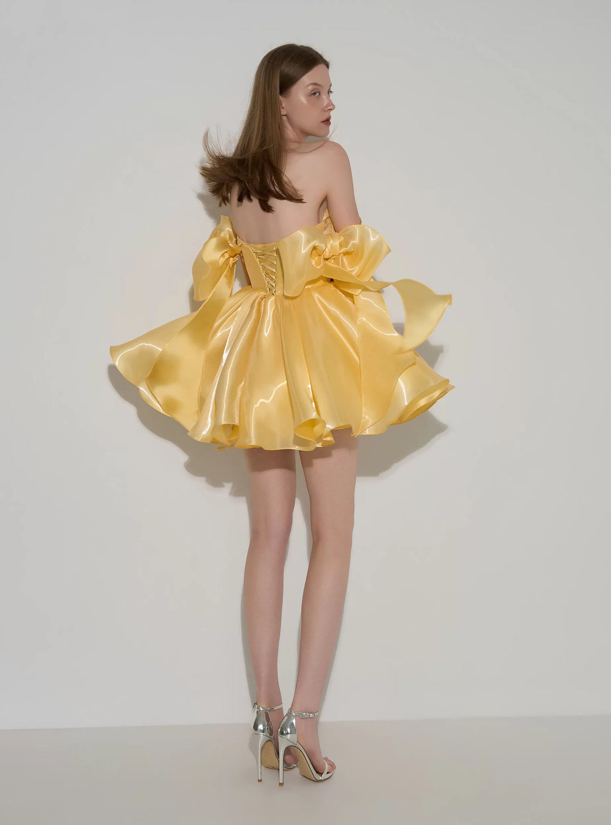 A woman modeling a strapless yellow satin dress with dramatic oversized bow details and a voluminous skirt, photographed against a plain light gray studio background.