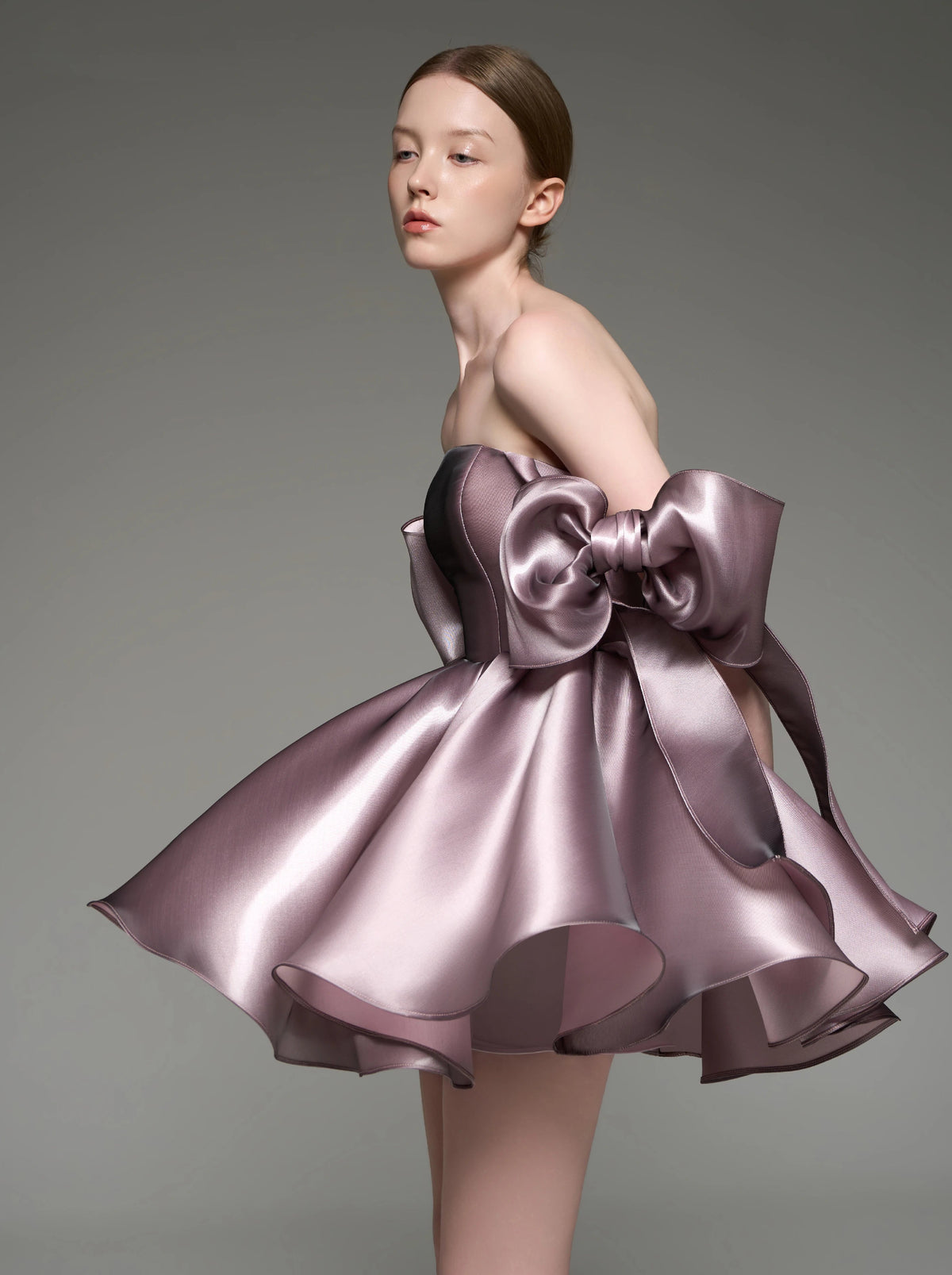 A model wearing a strapless formal dress made of shiny mauve-purple fabric with a large bow at the waist and a flared layered skirt, photographed against a plain gray studio background.