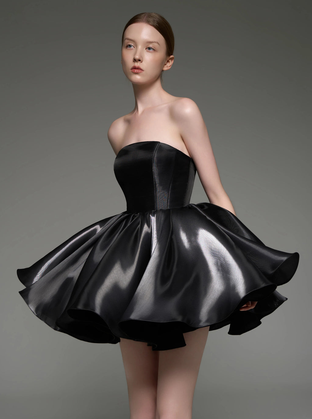 A model wearing a strapless black satin mini dress with a structured corset-style bodice and a flared, layered skirt, photographed against a neutral gray studio background.