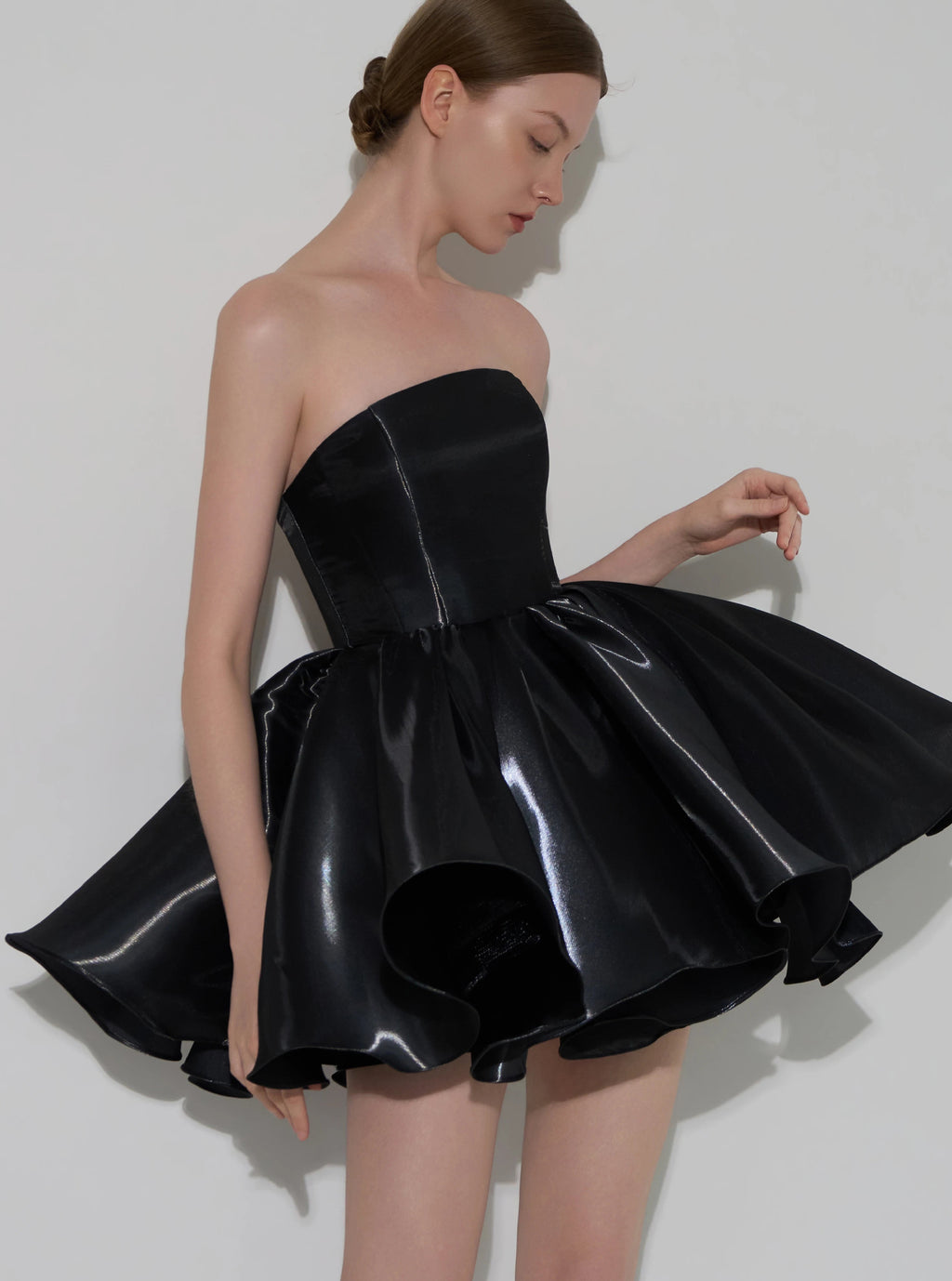 A model wearing a strapless black formal dress with a structured corset-style bodice and a flared, glossy skirt, posed against a light gray studio background.