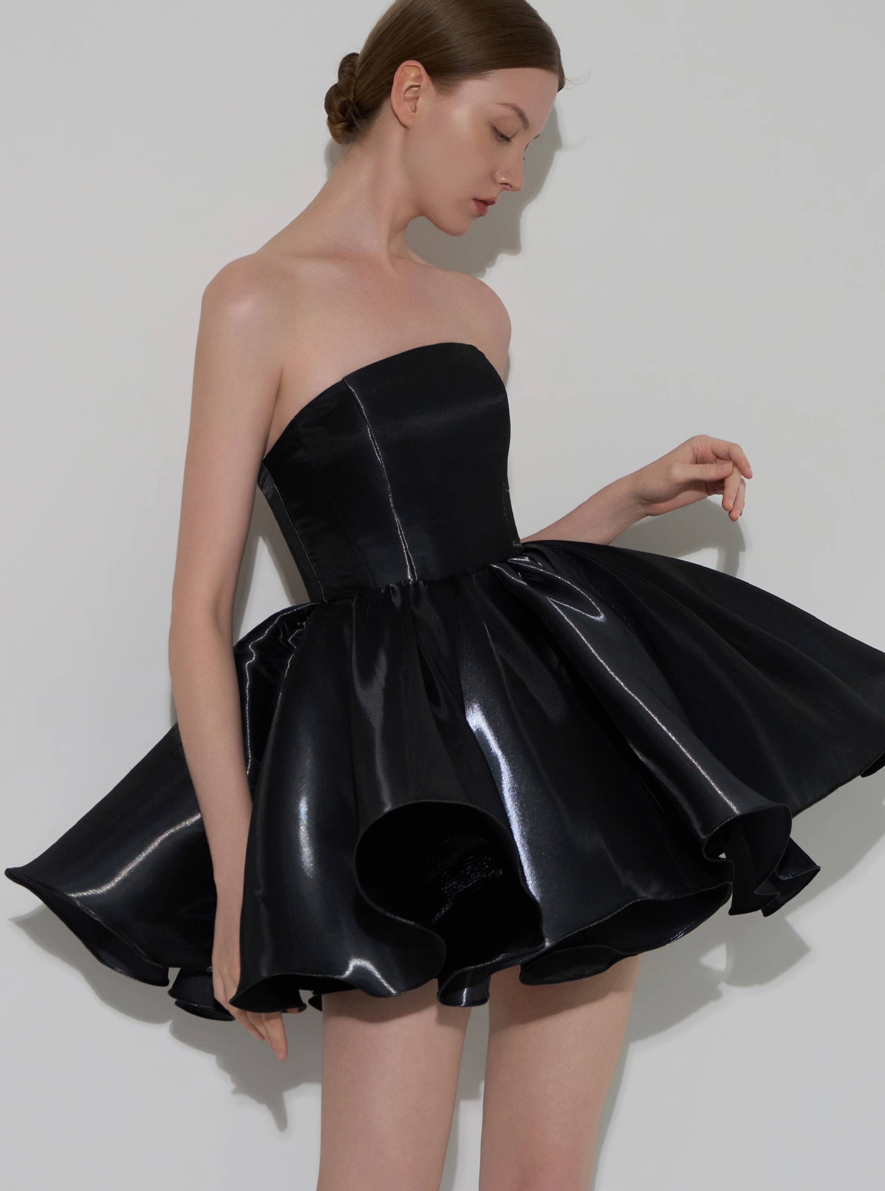 A model wearing a strapless black formal dress with a structured corset-style bodice and a flared, glossy skirt, posed against a light gray studio background.