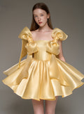 A model wearing a short, shiny pale yellow satin dress with oversized bow details on the shoulders, photographed against a plain gray studio background.