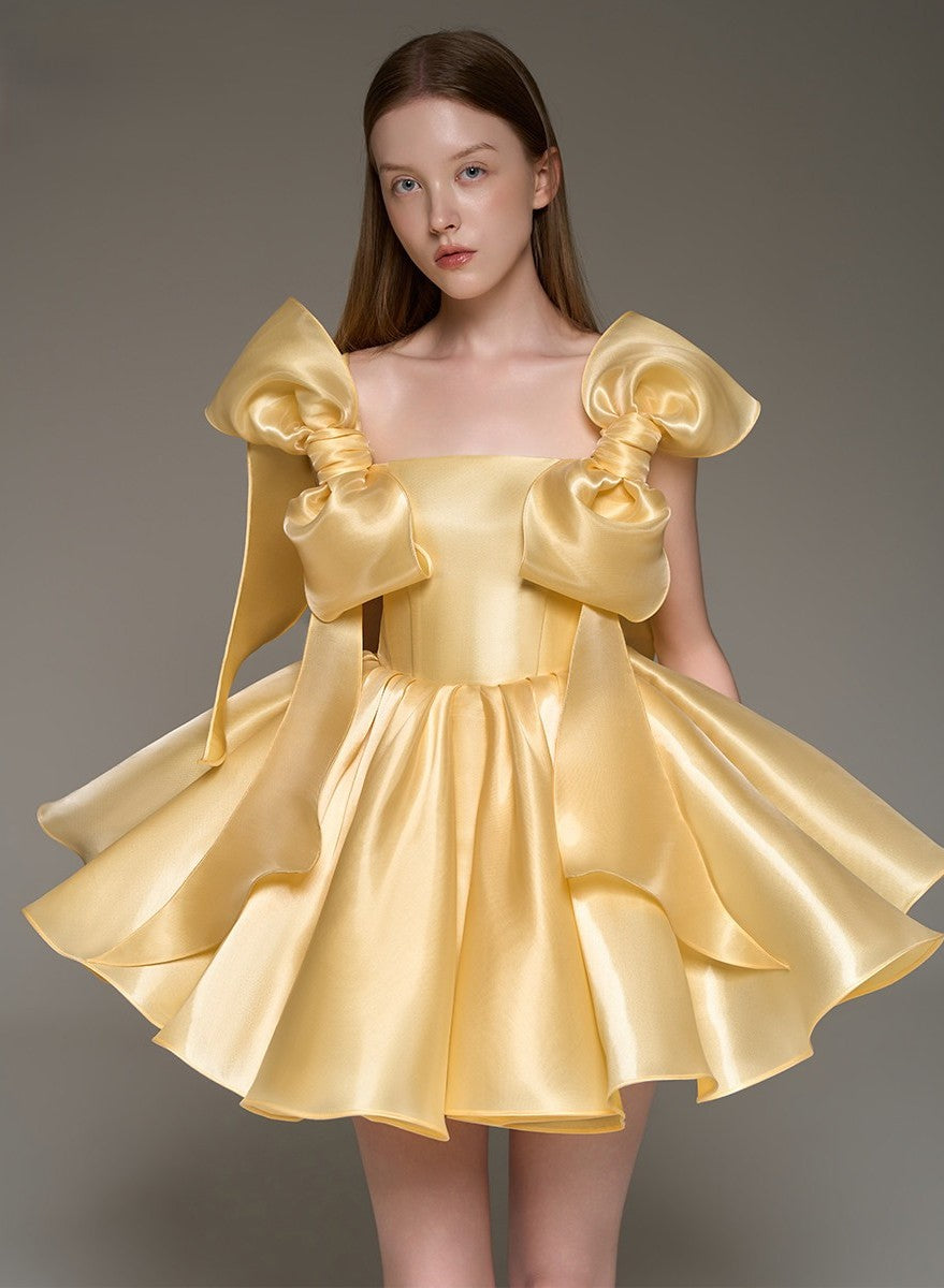 A model wearing a short, shiny pale yellow satin dress with oversized bow details on the shoulders, photographed against a plain gray studio background.