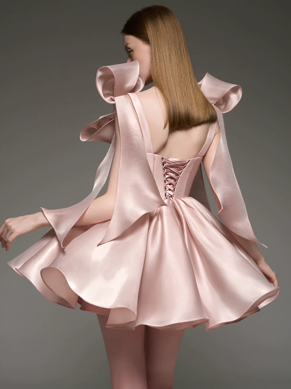 A woman shown from the back wearing a light blush pink satin formal dress with dramatic ruffled shoulder details, a lace-up open back, and a flared skirt, photographed against a dark gray studio background.