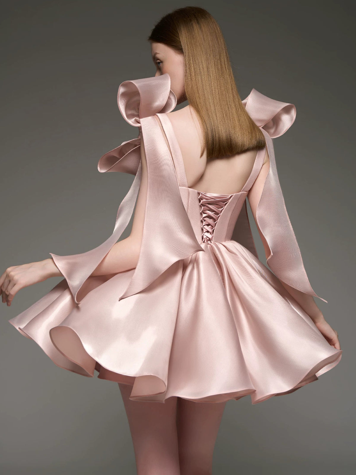 A woman shown from the back wearing a light blush pink satin formal dress with dramatic ruffled shoulder details, a lace-up open back, and a flared skirt, photographed against a dark gray studio background.