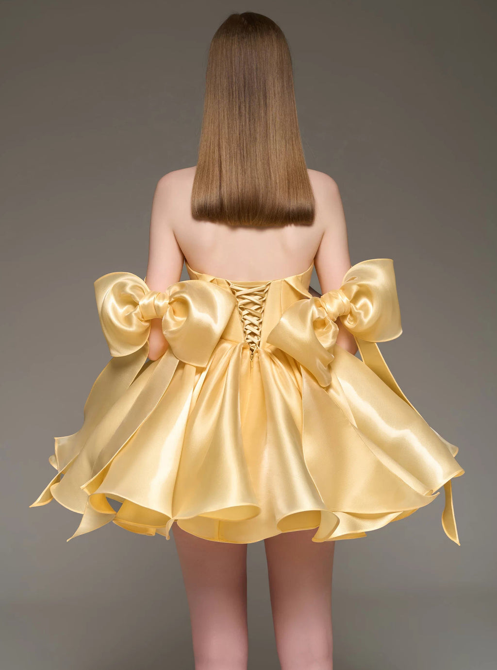 A woman shown from the back wearing a short formal dress made of shiny pale yellow satin. The dress has large bow details at the shoulders and a lace-up corset-style back.