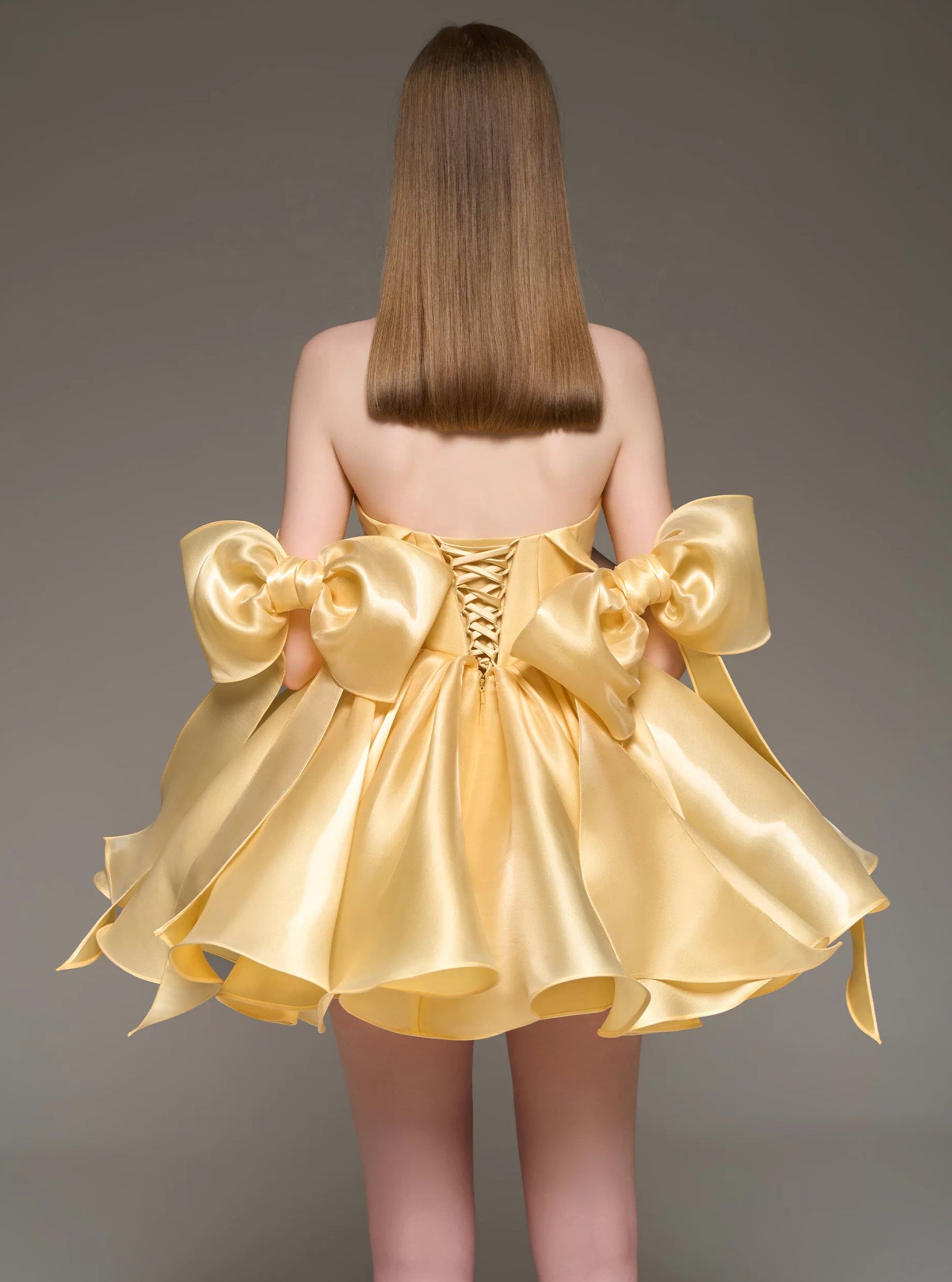 A woman shown from the back wearing a short formal dress made of shiny pale yellow satin. The dress has large bow details at the shoulders and a lace-up corset-style back.