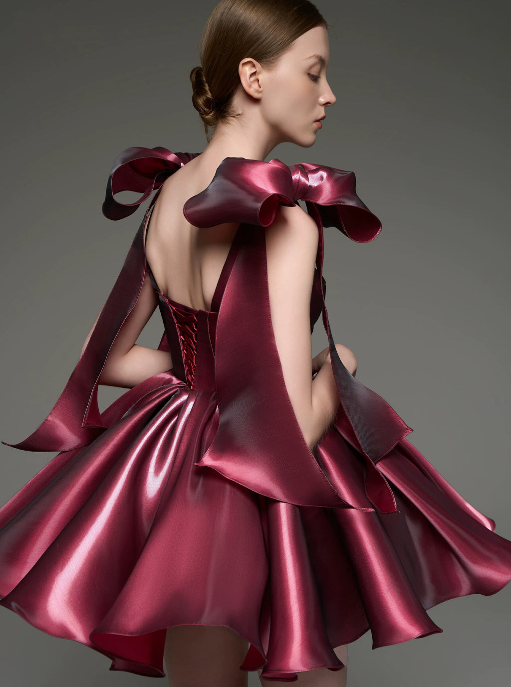 A model shown from the back wearing a metallic deep burgundy custom dress with dramatic oversized bow details on the shoulders and a structured flared skirt, set against a solid gray studio background.